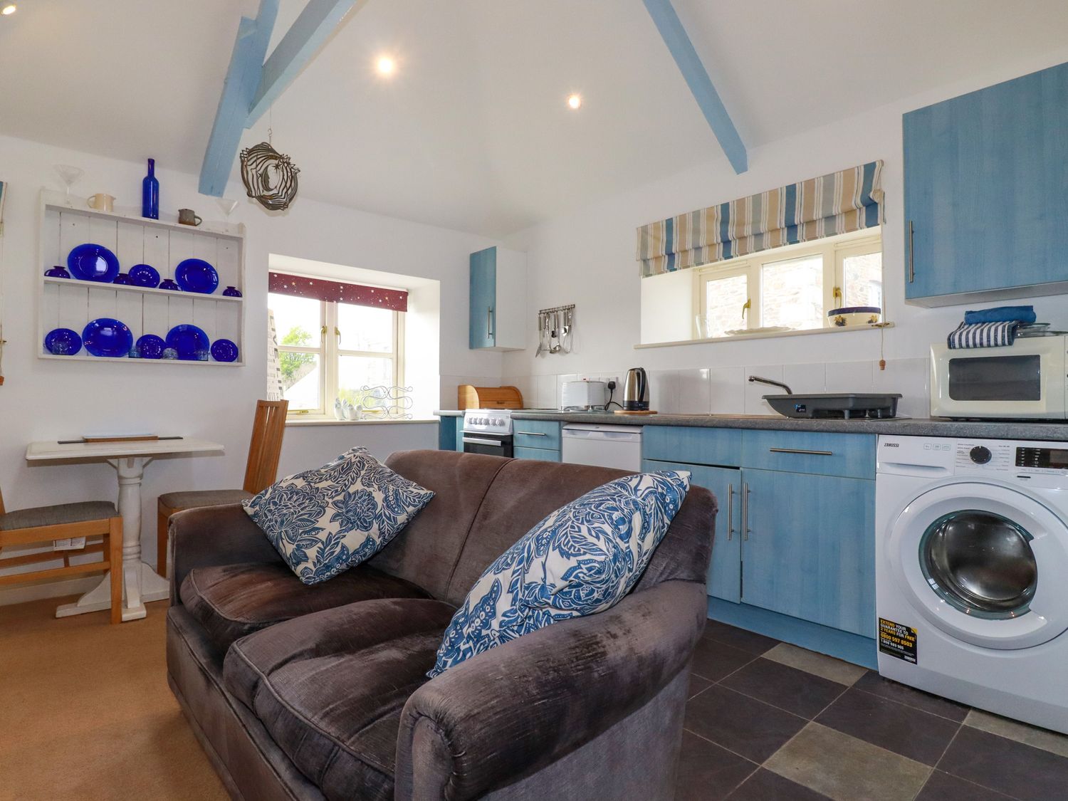 A kitchen with blue cabinets a gray sofa and blue plates on a wall shelf at Moonlight in St Erme near Truro