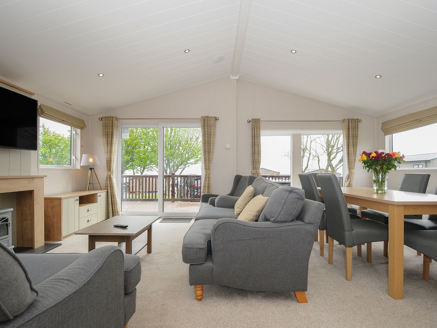 A living room with grey sofas, a wooden coffee table and a dining table with chairs and flowers on top at Lodge 64 at Riviera Bay Coastal Retreat in Brixham