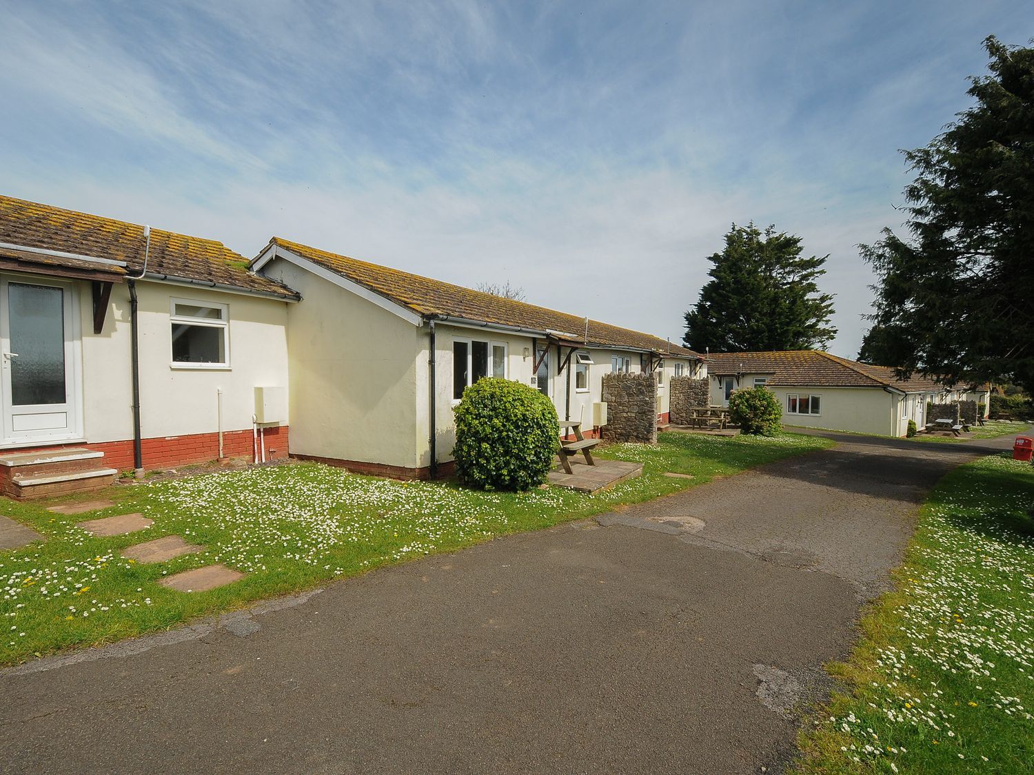 A path running past single-story holiday chalets with grassy areas and bushes at Chalet 38 at Landscove Holiday Park in Brixham