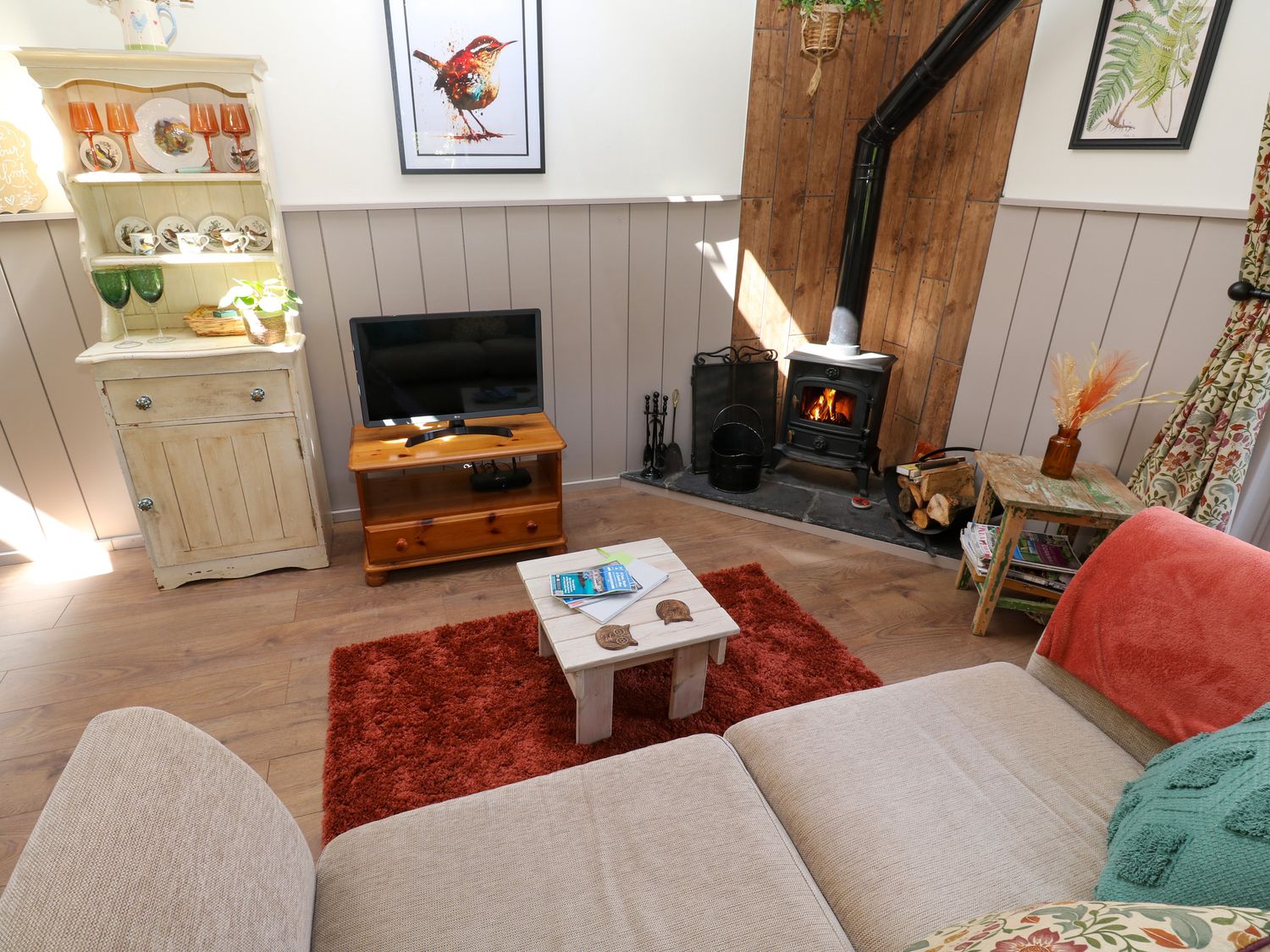 A living room with a wood burning stove a television on a wooden stand a beige sofa a small wooden table with books and coasters and a display cabinet with glassware at Little Wren Cottage in Marazion