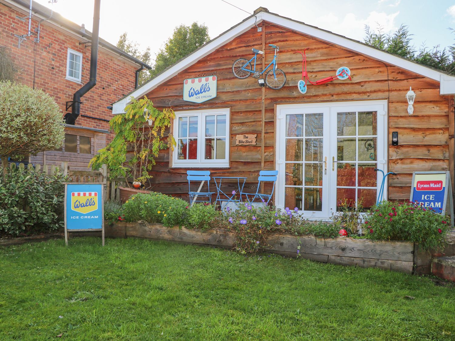 An outdoor area with a wooden shed featuring a small blue table and chairs a bicycle and scooter mounted on the wall and ice cream signs at The Bike Shed in Lockerley near West Tytherley