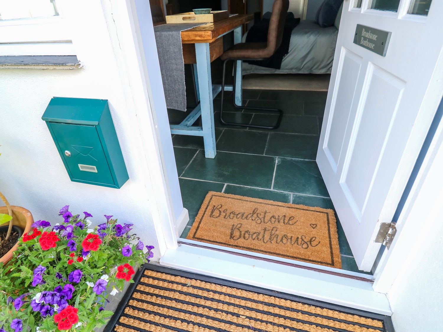 An open white door with a doormat that says Broadstone Boathouse flowers and a green mailbox outside at Broadstone Boathouse Dartmouth