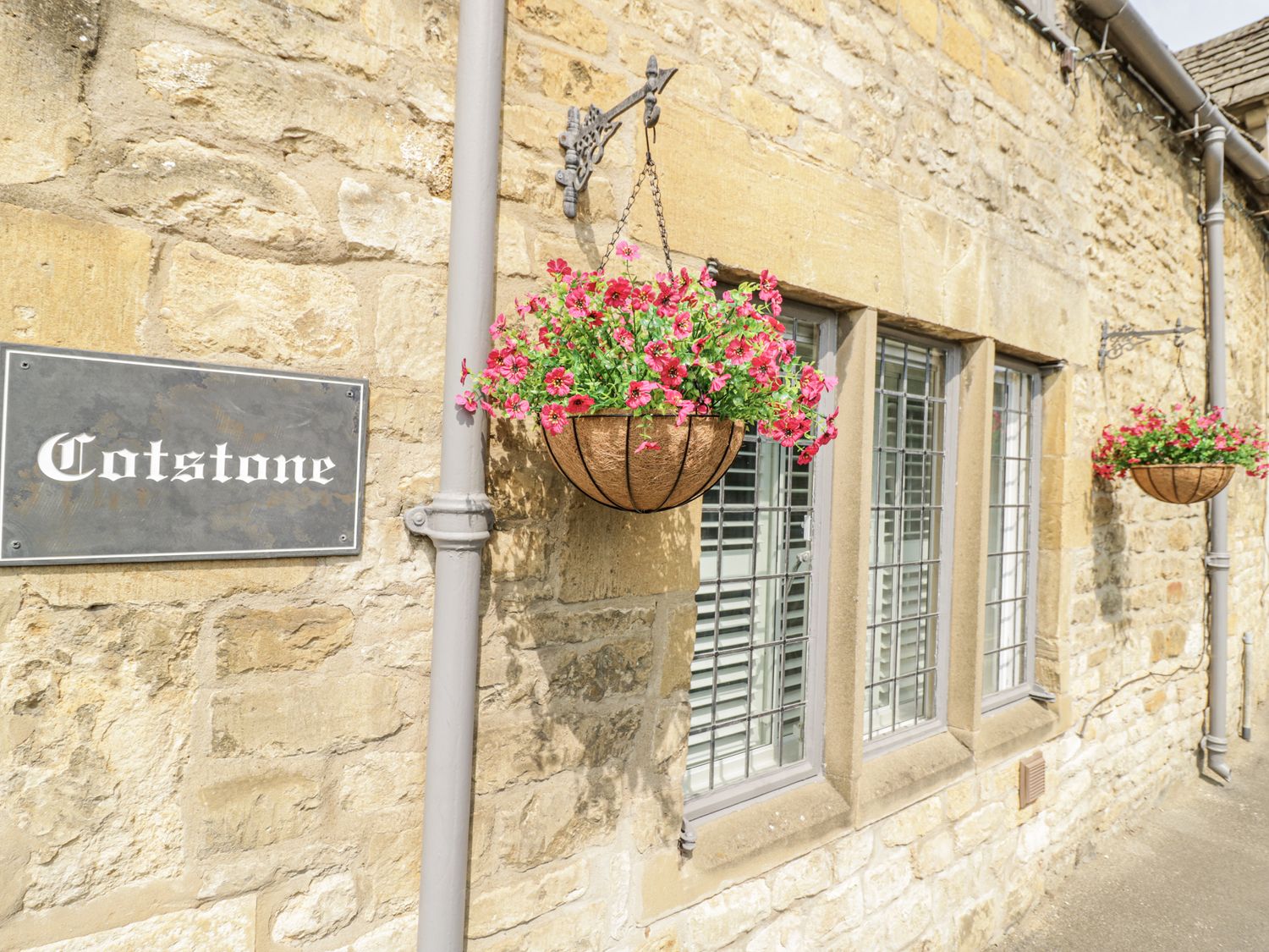A stone wall with hanging flower baskets and a sign at Cotstone Cottage in Chipping Campden