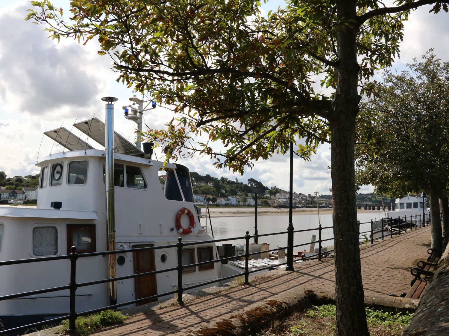 A boat docked beside a paved walkway with railings and trees along a waterfront at Woodland Roost in Parkham near Bideford
