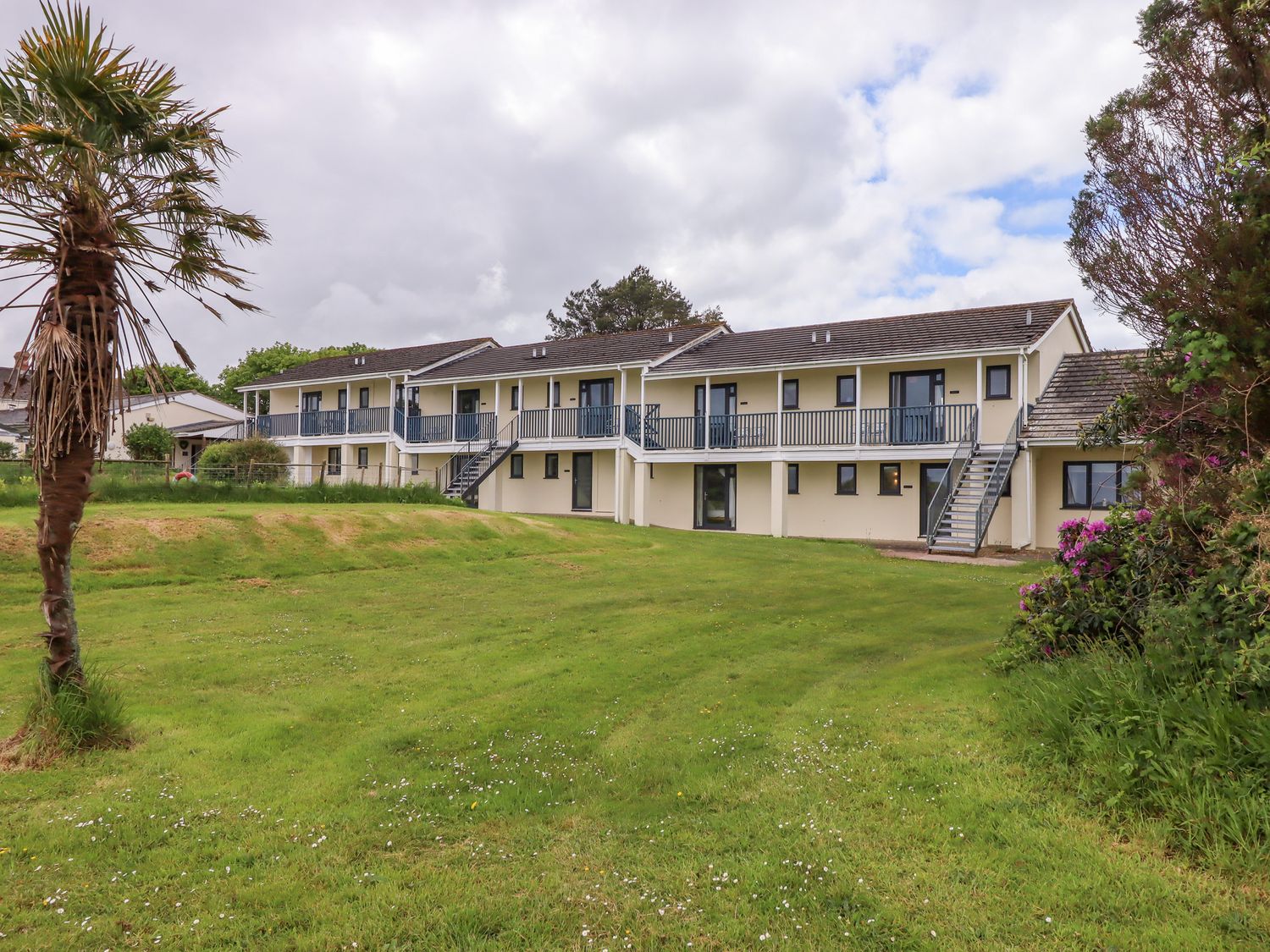 A two-story building with balconies and stairs overlooking a grassy lawn with a palm tree at Primrose at Moorhead Country Holidays Woolfardisworthy