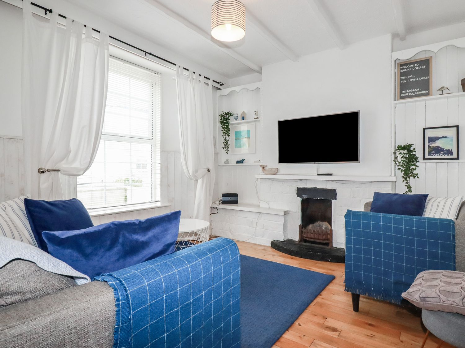 A living room with two sofas with blue cushions and throws a TV above a fireplace and a window with white curtains at Bluejay in Newquay