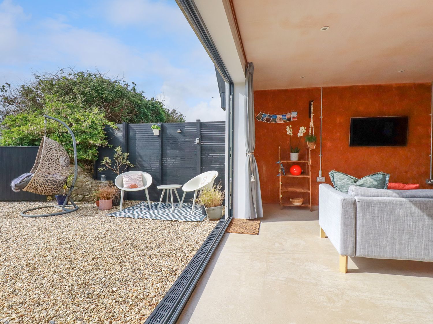 An open living area with a gray sofa and a TV mounted on a red wall next to an outdoor seating area with two chairs a hanging chair and plants at Gwynn lodge in Newquay