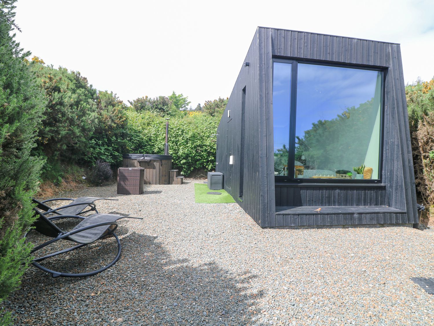 An outdoor area with two lounge chairs a hot tub and a black modern cabin surrounded by shrubbery at Stargazer Tara Hill near Ballymoney County Wexford