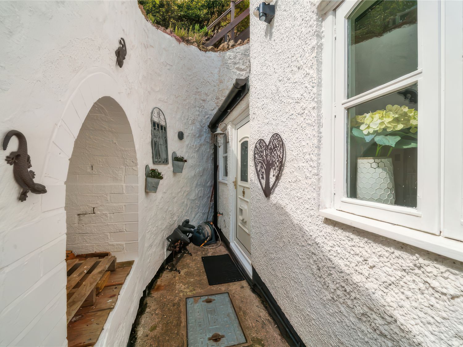 A narrow outdoor passageway with white textured walls a white door a window with a vase of flowers and decorative metal wall hangings at Holly Cottage in Great Malvern