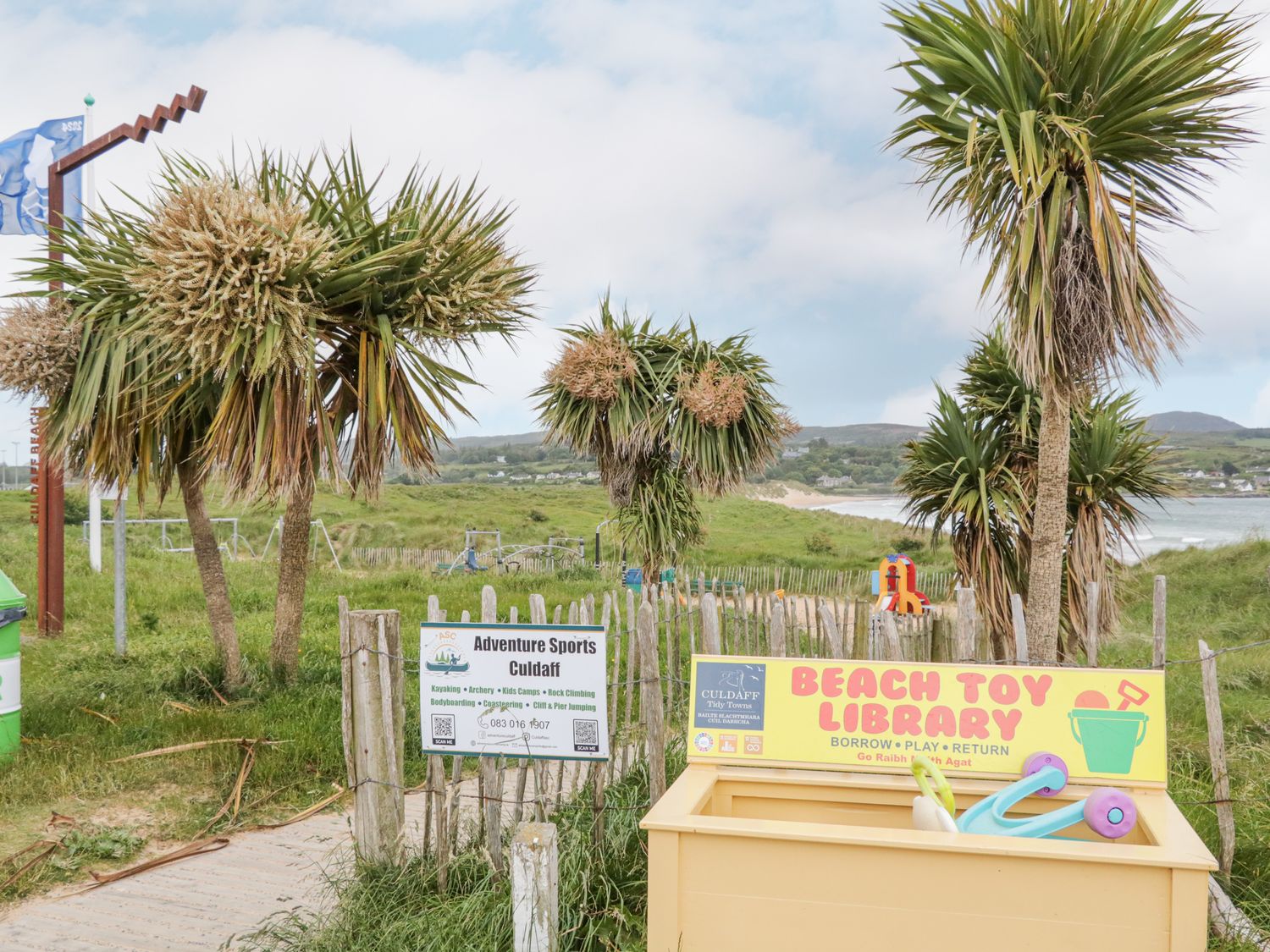 An outdoor area with palm trees a wooden fence signs for adventure sports and a beach toy library at Stoneywood Culdaff County Donegal