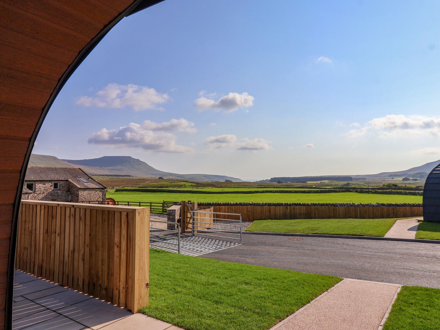 A view of green fields and distant hills from a curved wooden structure with a fenced yard at Wren in Ingleton
