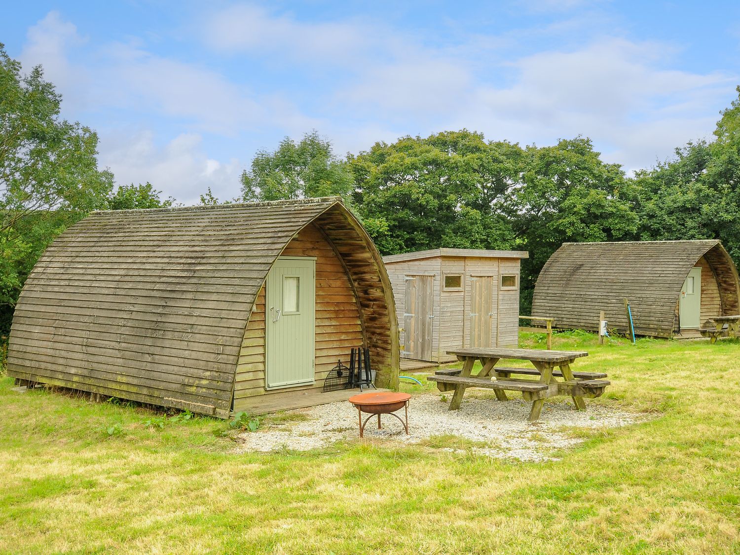 Two wooden camping pods with green doors near a wooden picnic bench and fire pit on grass at Rabbit Warren at Penbugle Organic Farm in St Keyne near Duloe