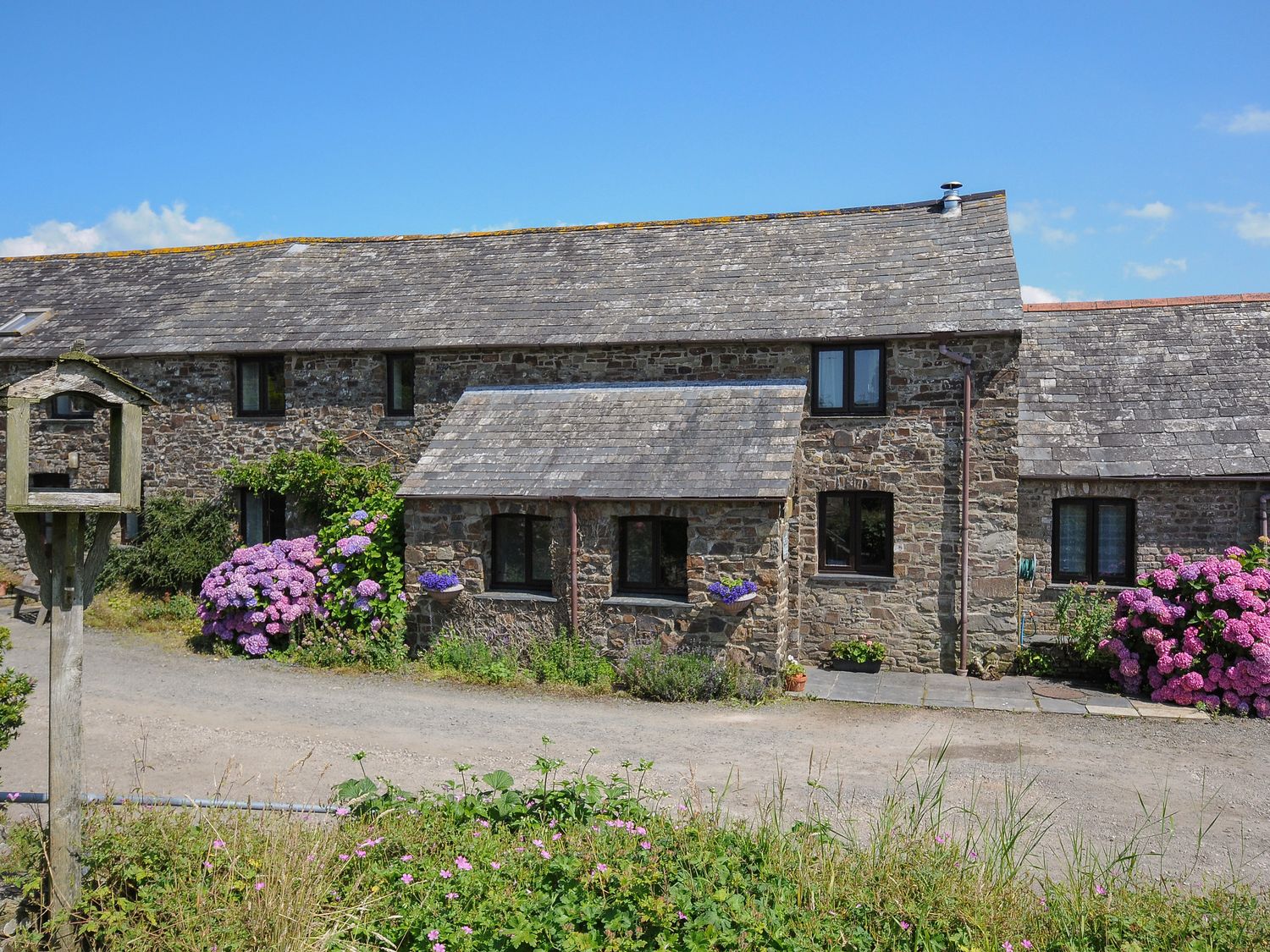 A stone house with multiple windows flowers and plants along the front and a clear sky at West Lee in Bude