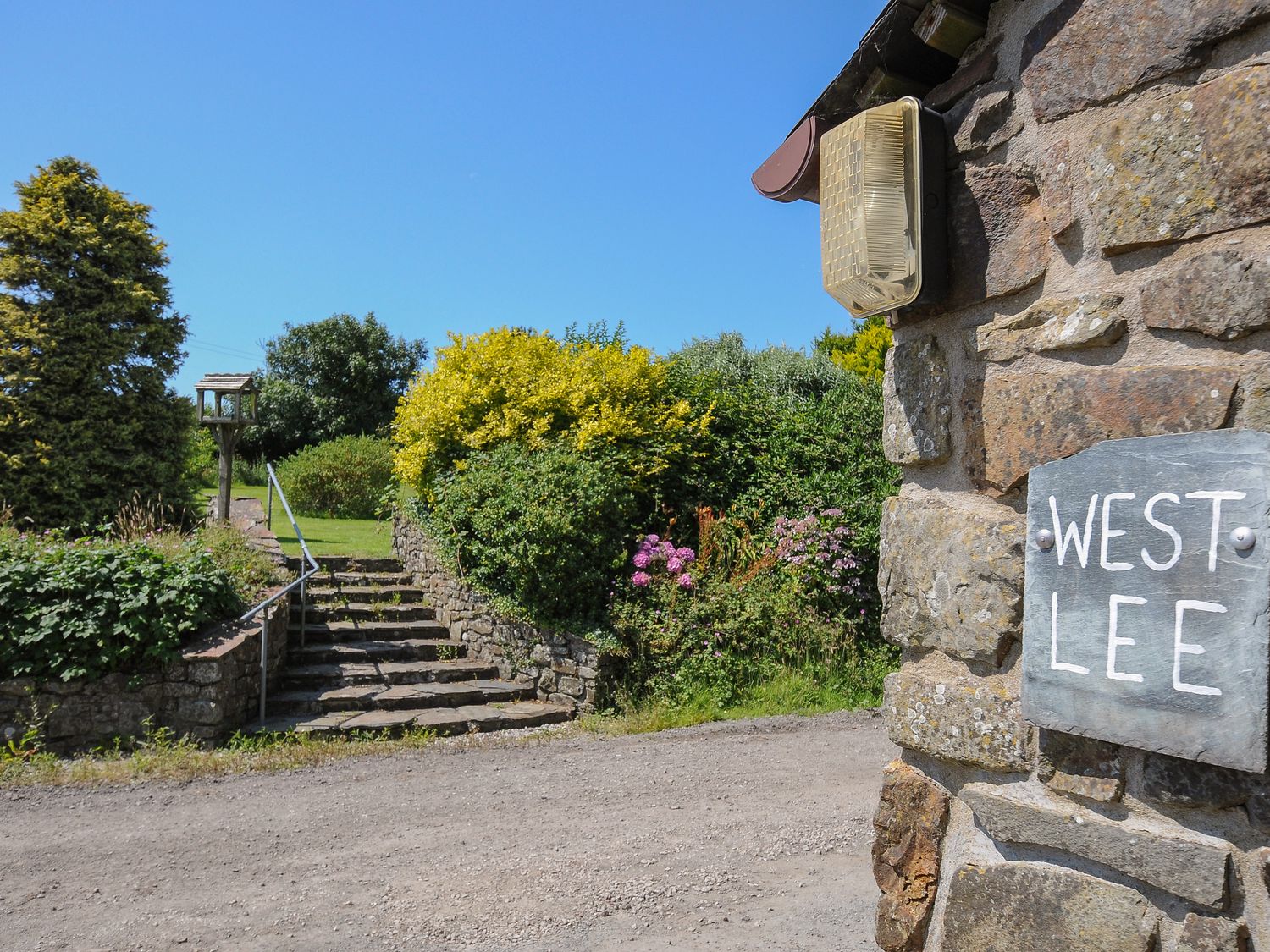 Stone steps leading to a garden with bushes and trees and a stone wall with a West Lee sign in Bude