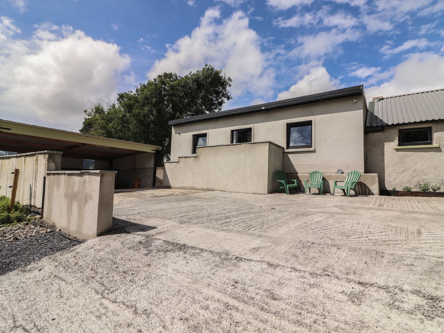 An outdoor area with a beige building a covered parking area and three green chairs against the building at Hogans Cottage Cullenagh near Ennistymon County Clare