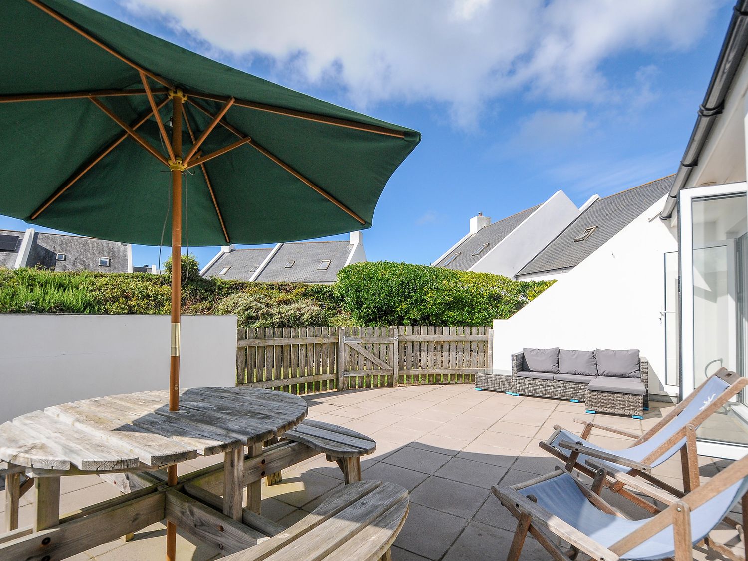 An outdoor patio with a wooden picnic table and umbrella two lounge chairs and a sofa with cushions at Bryher in Trebetherick near Polzeath