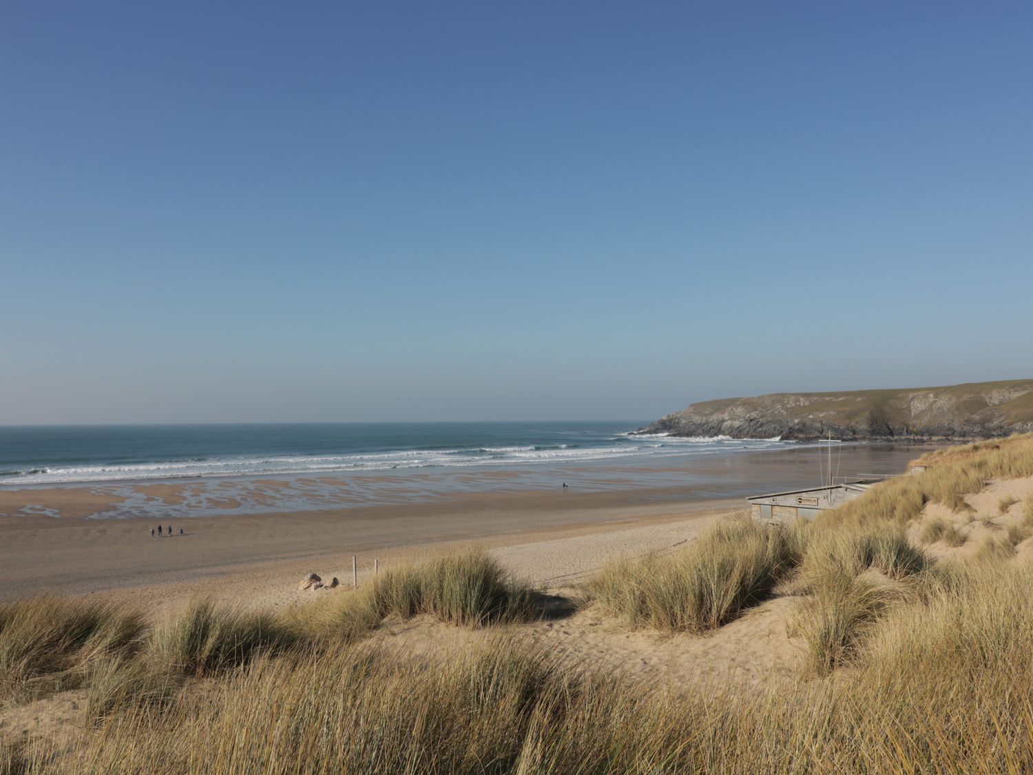 A sandy beach with waves and grassy dunes with cliffs in the background near Horseshoe Cottage Near Holywell Bay