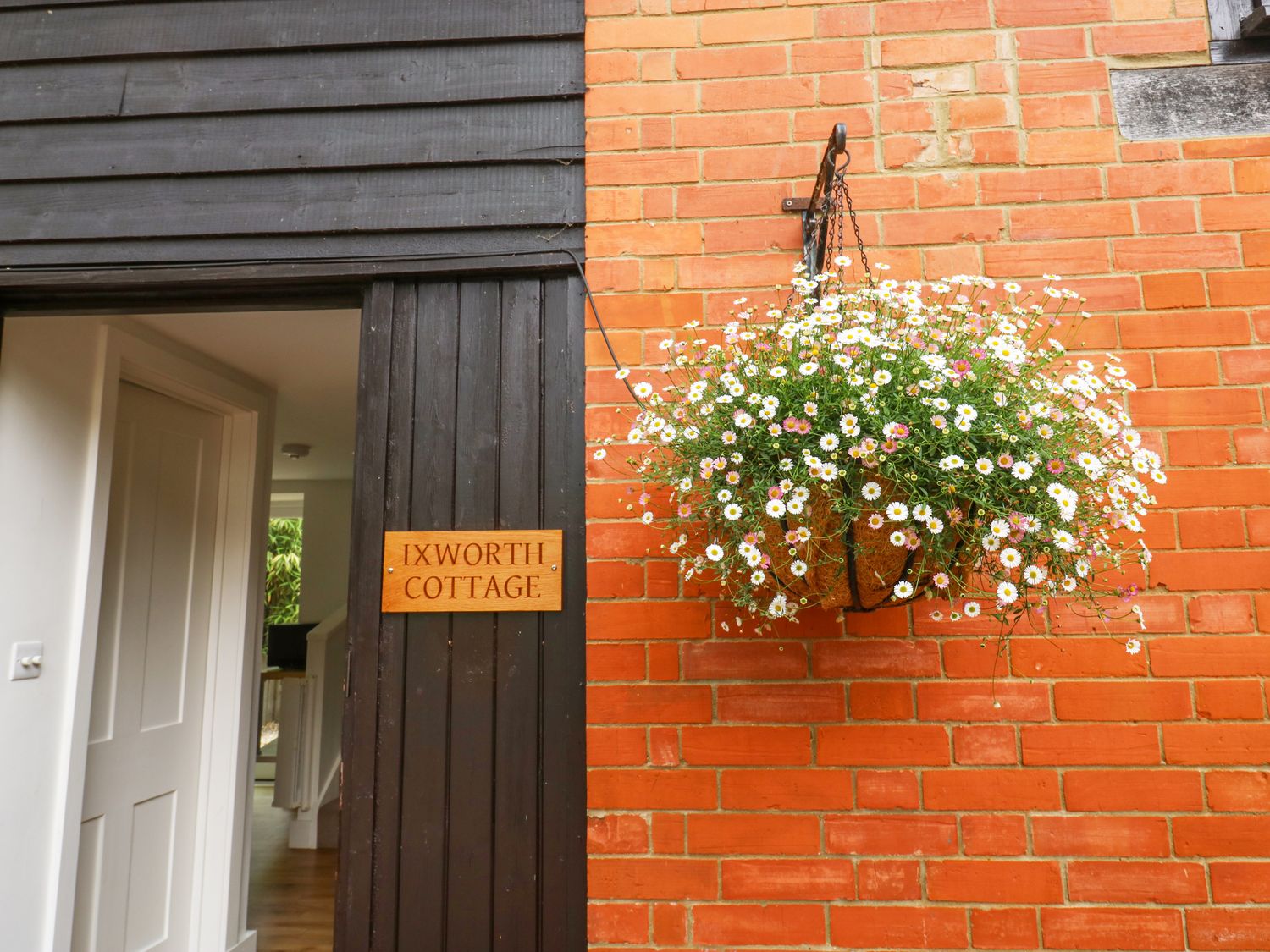 An open door with a wooden sign reading Ixworth Cottage and a hanging basket of flowers on a brick wall at Ixworth Cottage in Little Woodford near Ottery St Mary