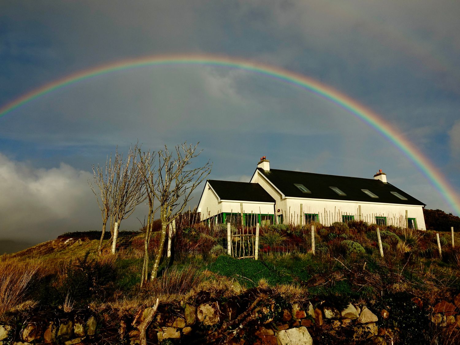 A house on a hill with leafless trees and a rainbow in the sky at Lighthouse Hill in Tiernaur near Mulranny