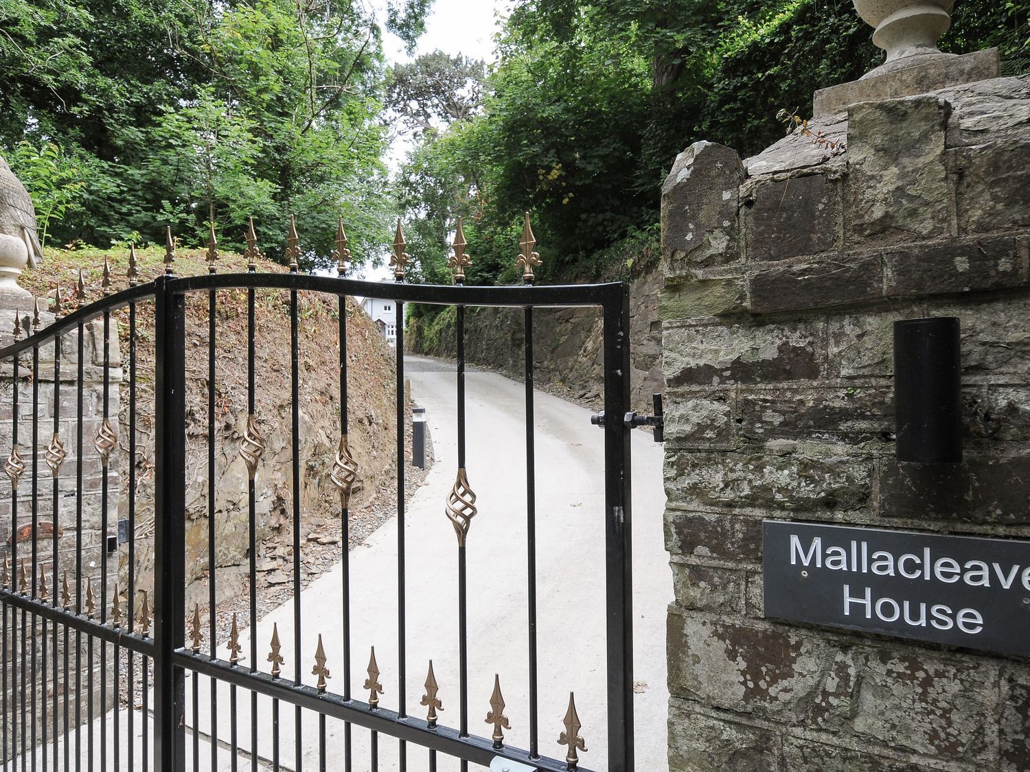 A black metal gate with gold spikes next to a stone wall with a sign reading Mallacleave House surrounded by trees and a driveway
