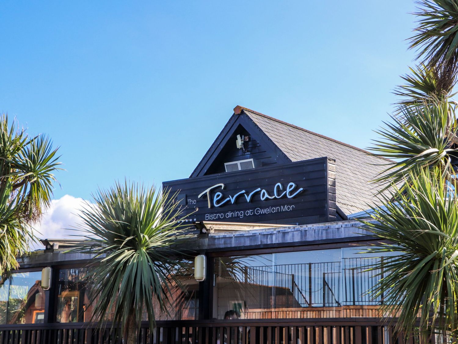 Exterior view of a bistro dining building with a sign reading The Terrace Bistro dining at Gwelan Mor with palm trees around Trevaunance in Portreath