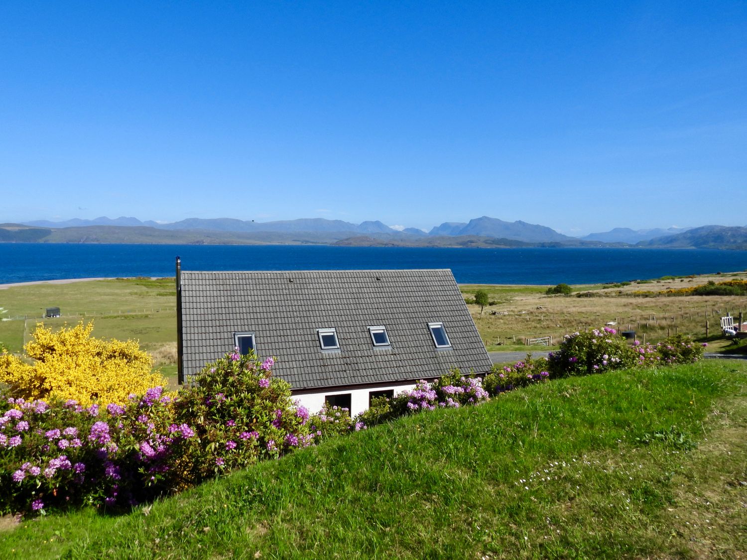 A house with a gray roof surrounded by flowering shrubs overlooking a body of water with mountains in the distance at Calmdale in Inverasdale near Poolewe