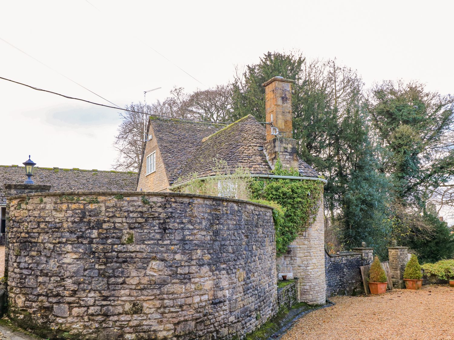 A stone house with a curved stone wall and trees in the background at The Elm Coach House in Chipping Norton