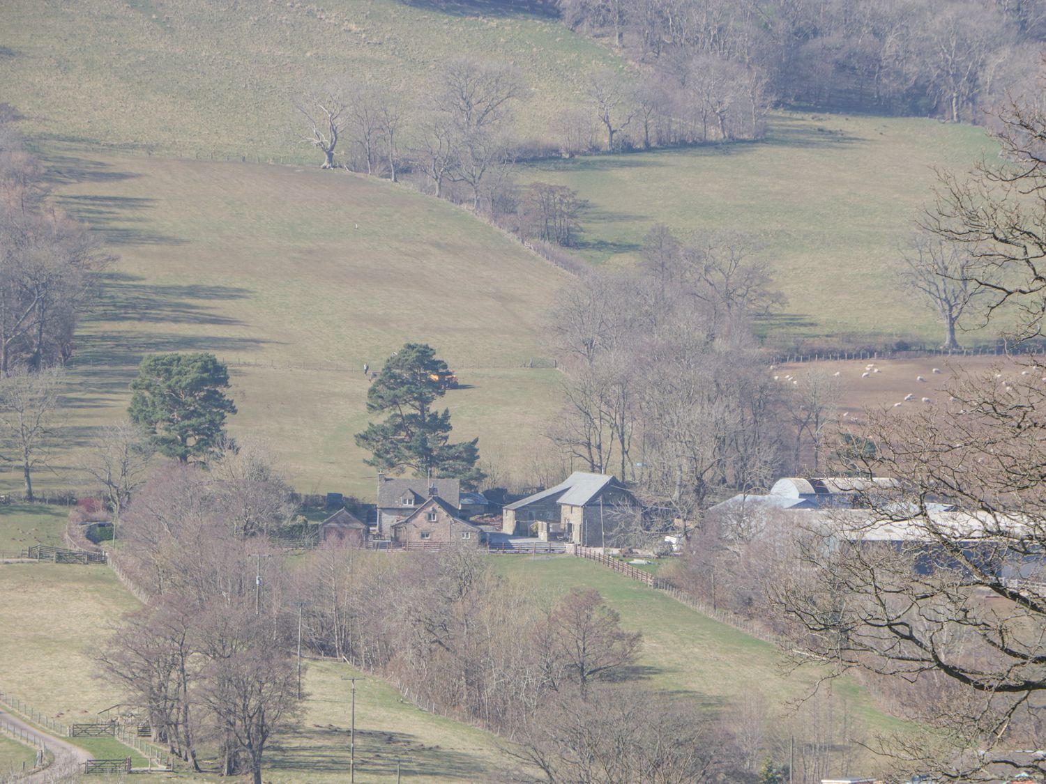 A rural farmstead with several buildings and trees surrounded by fields and fences at White Haywood Barn in Craswall