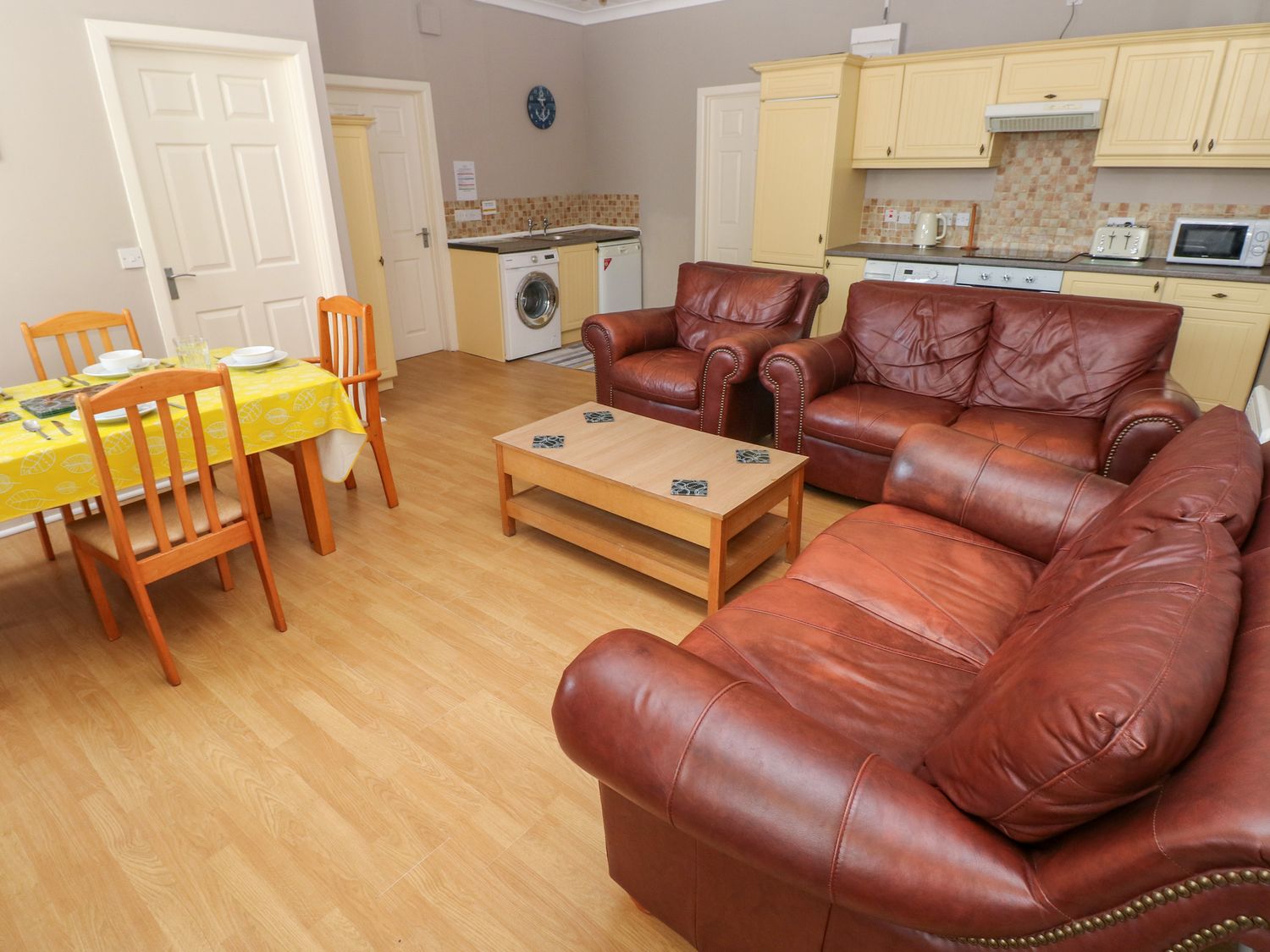 A living area with brown leather sofas a wooden coffee table a dining table with chairs and a kitchen with cream cabinets at Hilton Snug in Tenby