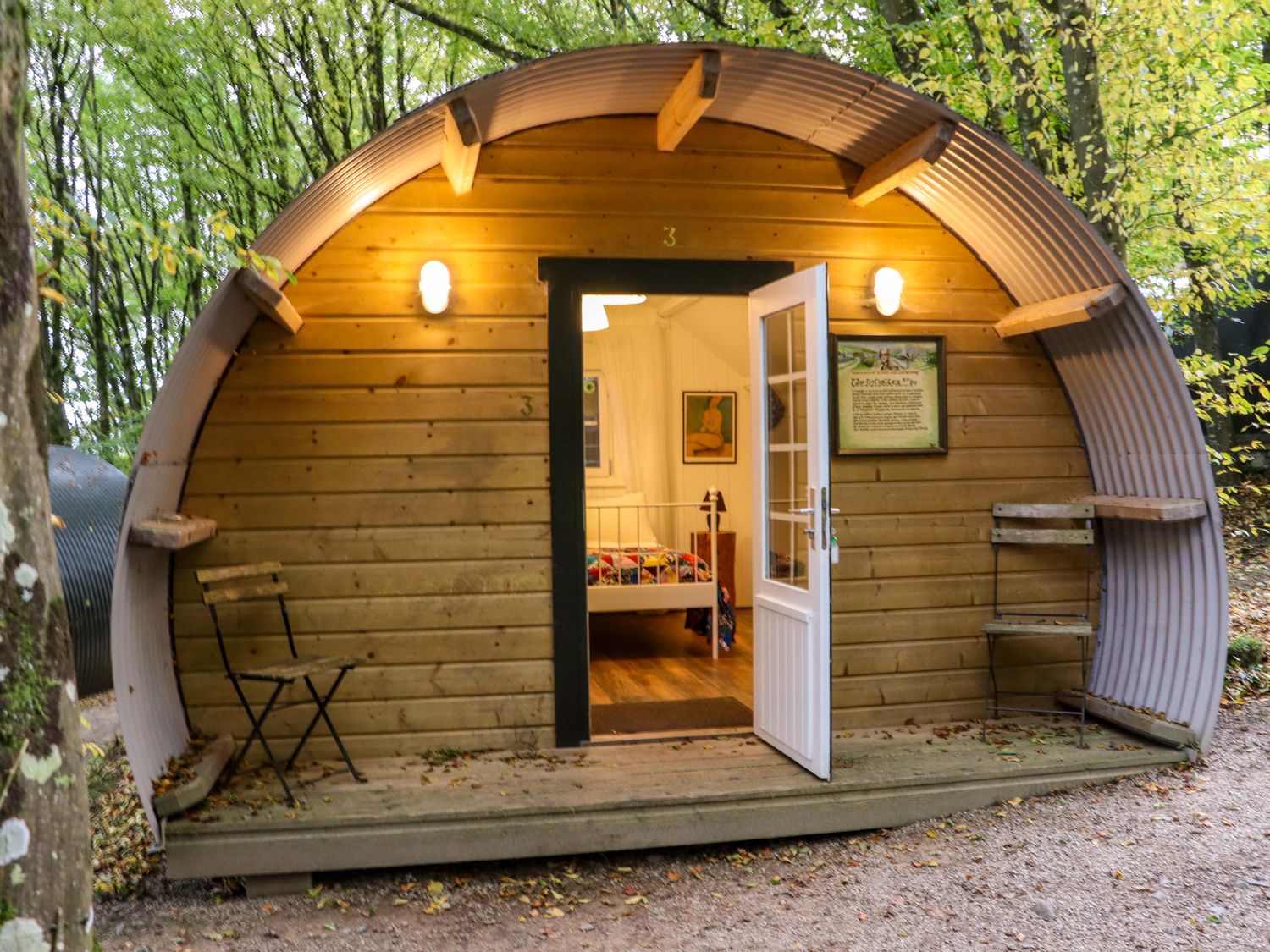 A curved wooden pod with an open door and two chairs outside in a forest at Ballybeg Pod 1 Ballybeg near Tinahely County Wicklow