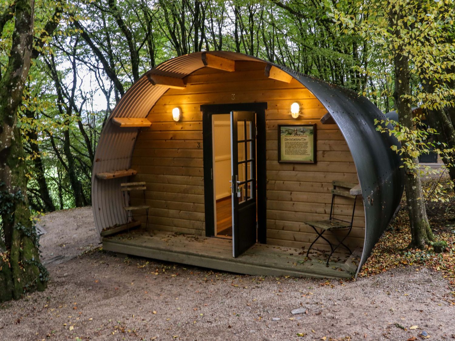 A wooden pod with curved metal roof in a forest with a door open and two metal chairs outside at Ballybeg Pod 7 Ballybeg near Tinahely County Wicklow