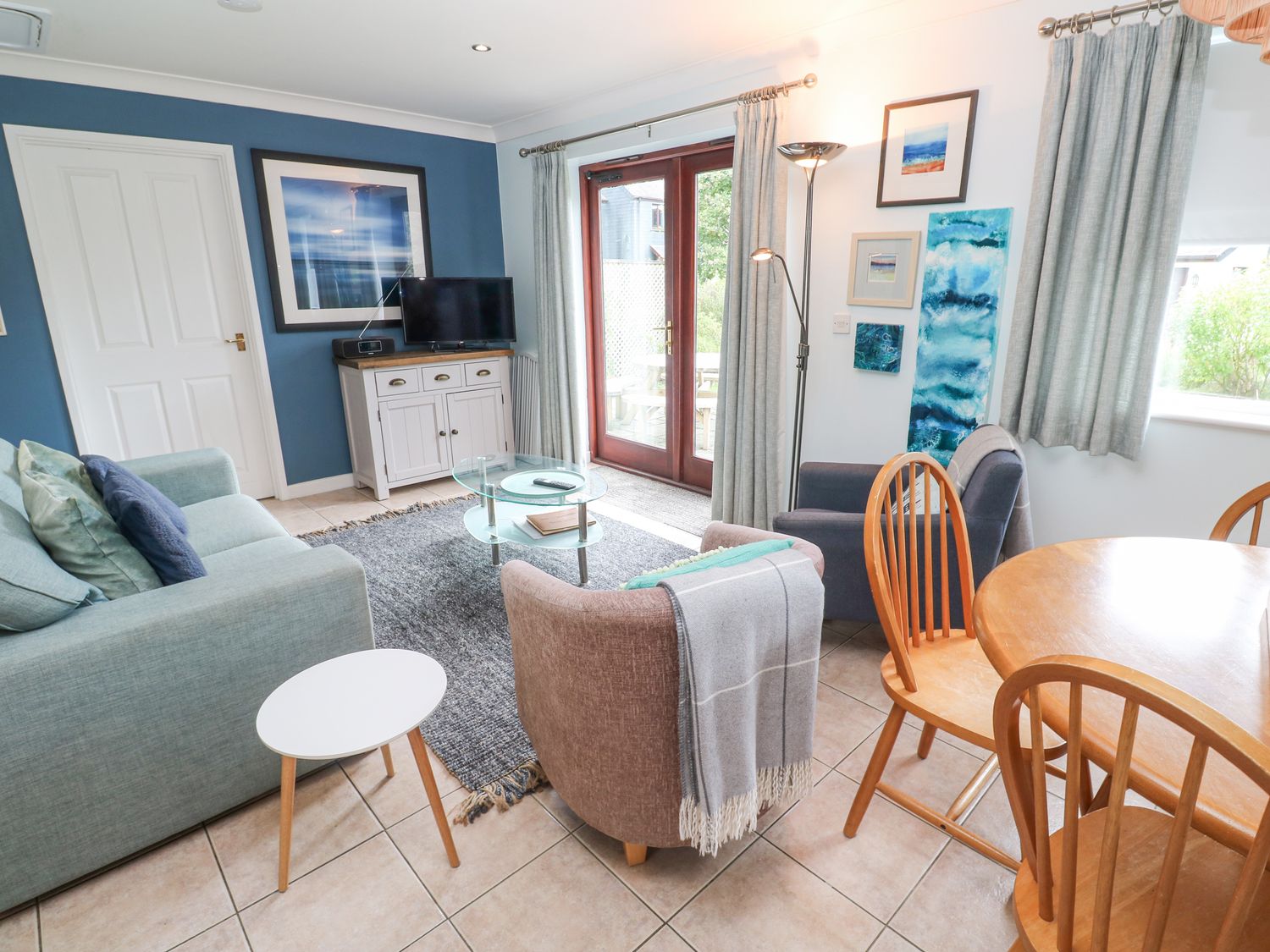 A living room with a blue sofa gray rug glass coffee table and wooden dining table with chairs at Chy Lowena in Goldenbank near Budock Water