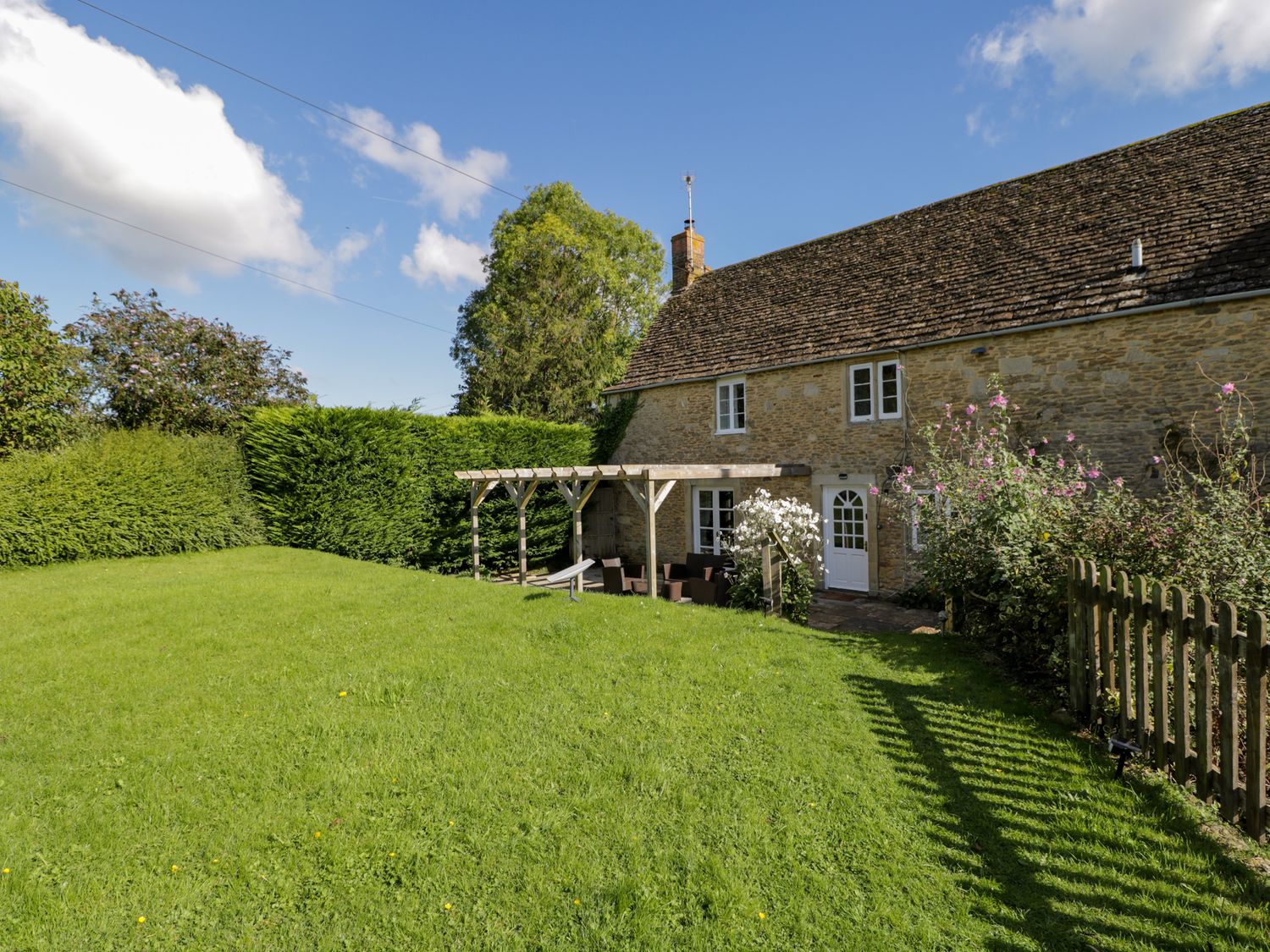 A lawn with wooden pergola and seating area next to a stone house with white doors and windows at Little Wick in Lacock