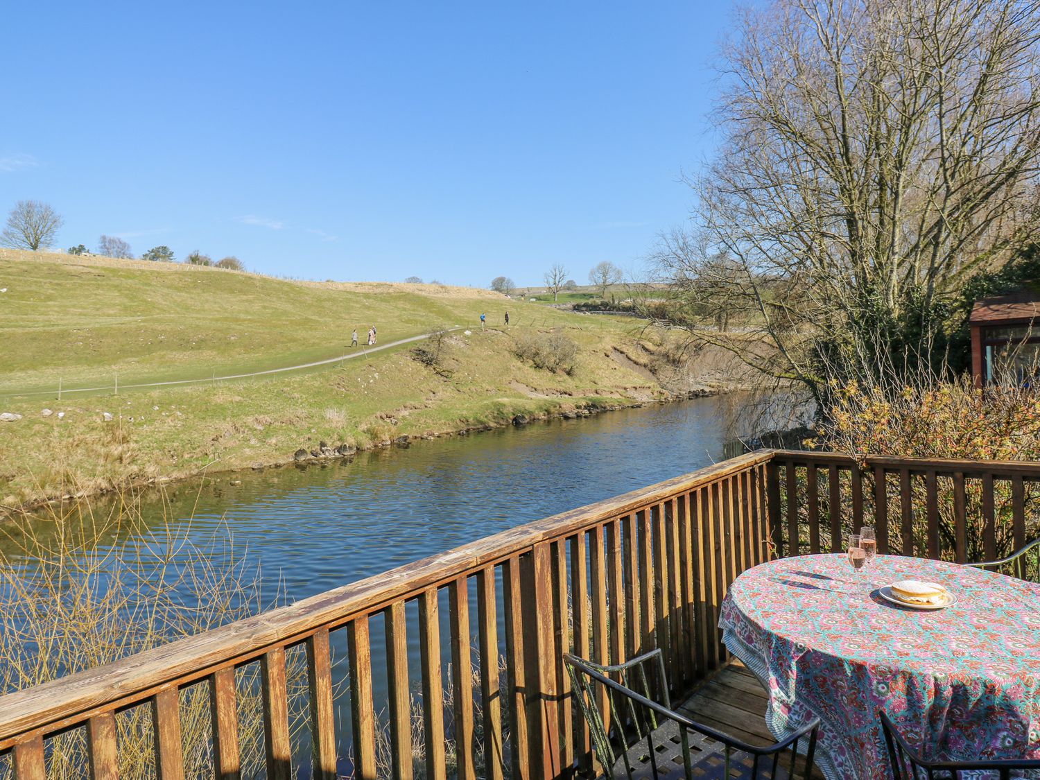 A wooden balcony overlooking a river with a table covered by a patterned cloth and two glasses at Kirk Yett Cottage in Linton Falls