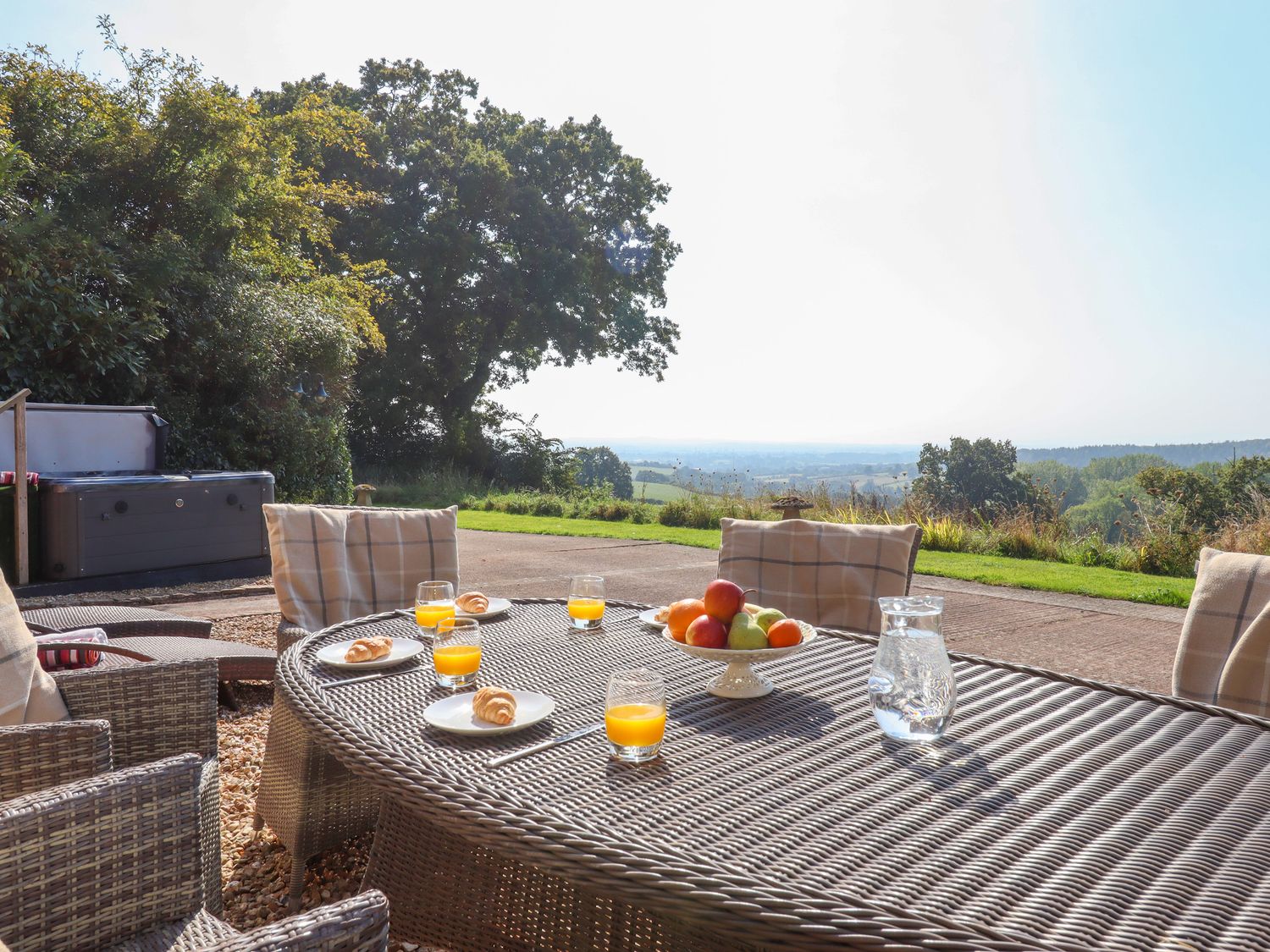 An outdoor patio with a wicker table and chairs glasses of orange juice plates with croissants a bowl of fruit and a pitcher of water at Sherway Coach House in Clyst Hydon near Bradninch