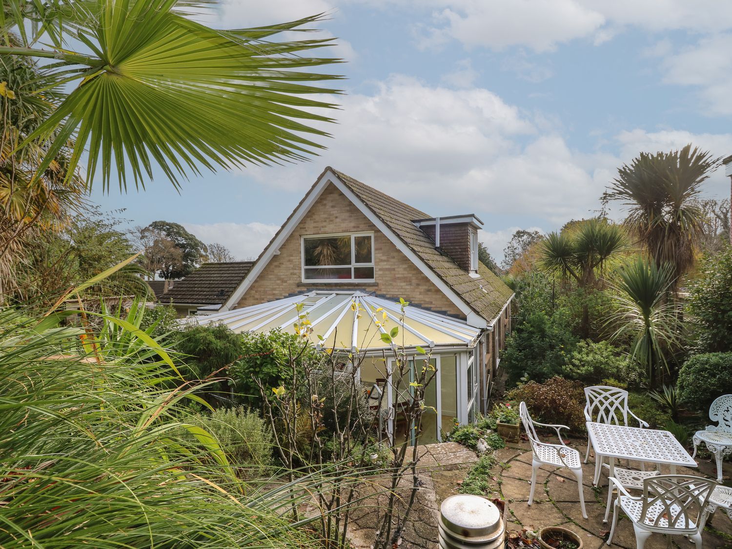 A garden with white patio furniture surrounded by plants and a house with a conservatory at Tall Timbers in Ventnor