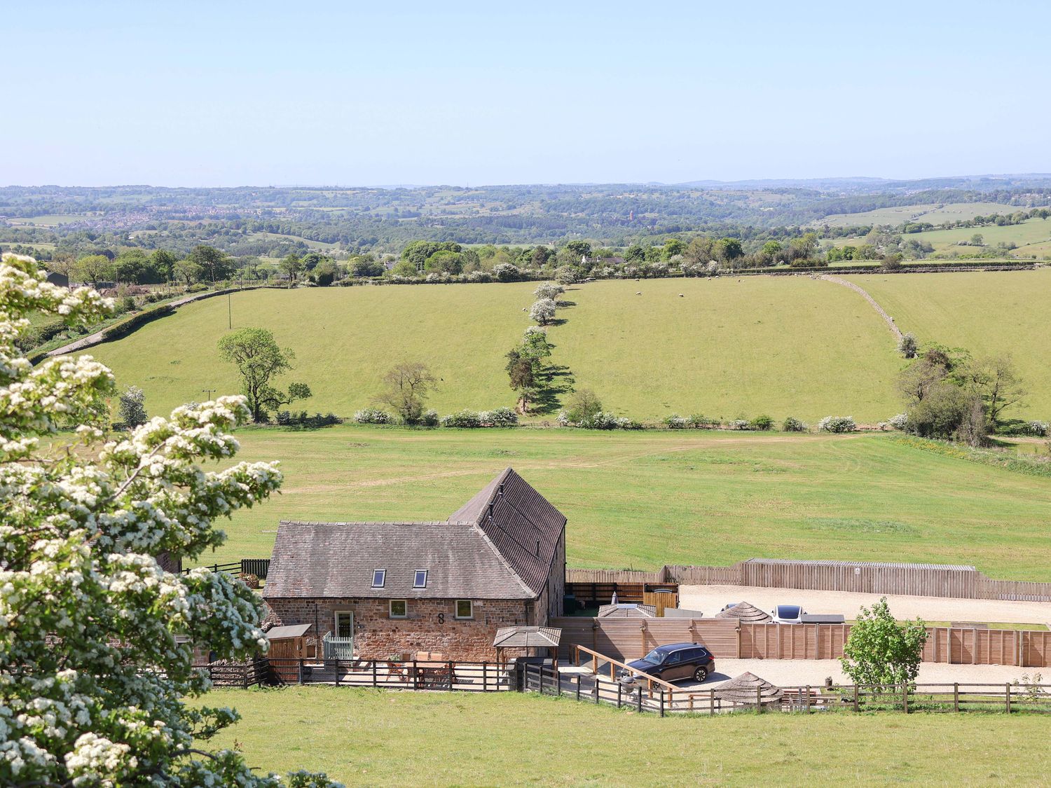 A barn and a car parked beside a fenced area with green fields and trees in the background at Parlour Barn in Leek