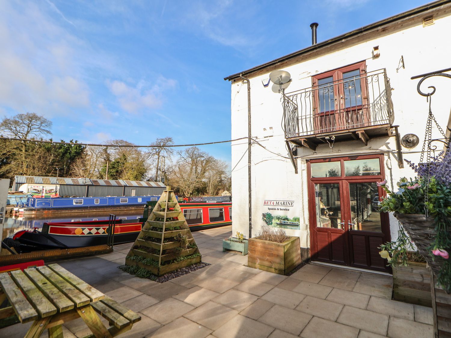 An outdoor patio area with wooden picnic table wooden planters and a white building next to a canal with narrowboats at Quayside Apartment in Nantwich