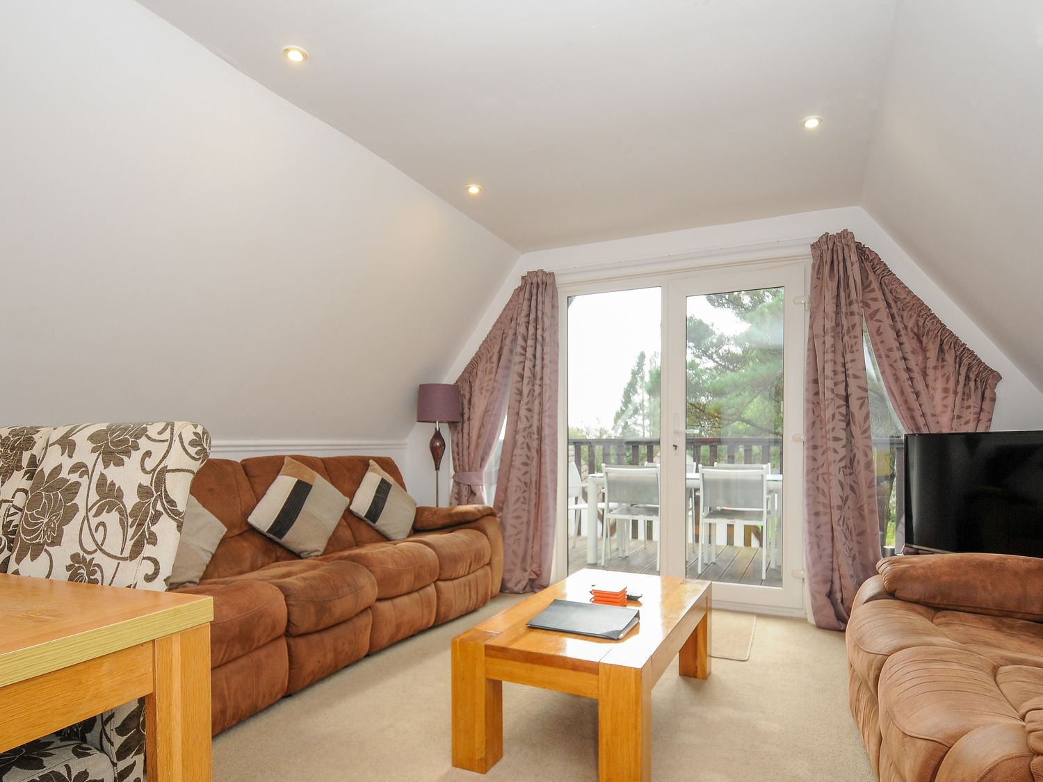 A living room with brown sofas wooden coffee table dining table and chairs visible through glass doors at Valley Lodge 26 in St Ann's Chapel near Callington