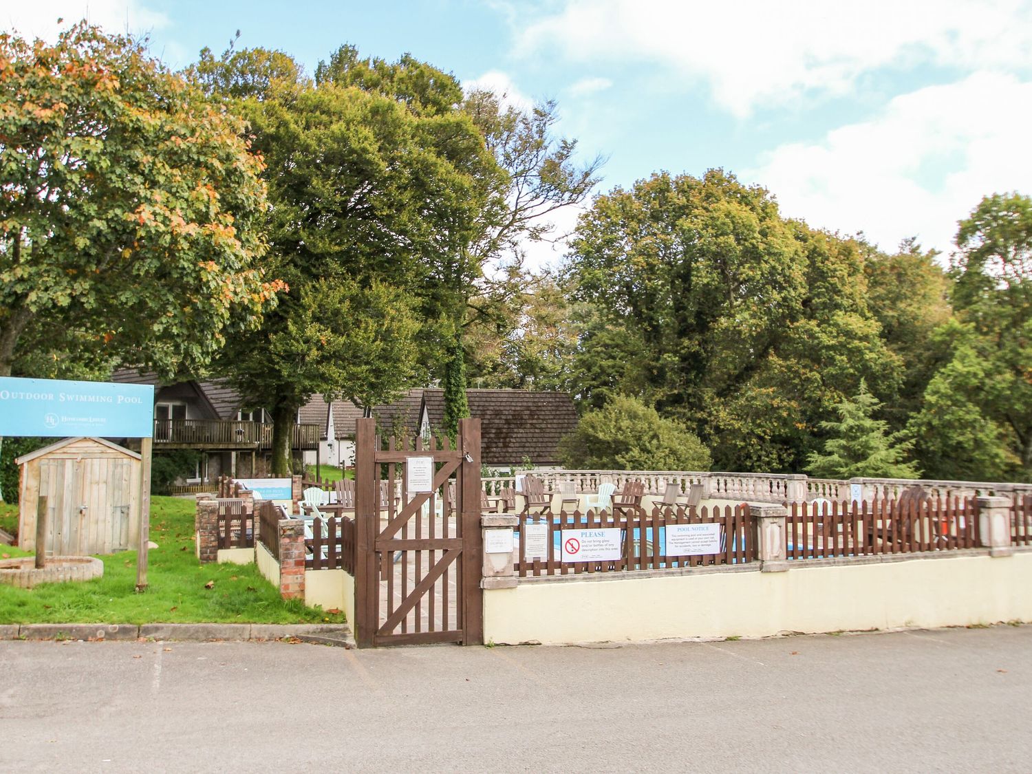 An outdoor swimming pool surrounded by a wooden fence with trees and buildings in the background at Valley Lodge 26 in St Ann's Chapel near Callington