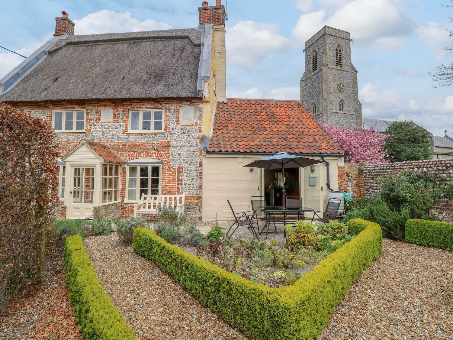 A garden with trimmed hedges and outdoor seating in front of a cottage with a thatched roof and a church tower in the background at Thatch Cottage in Trunch