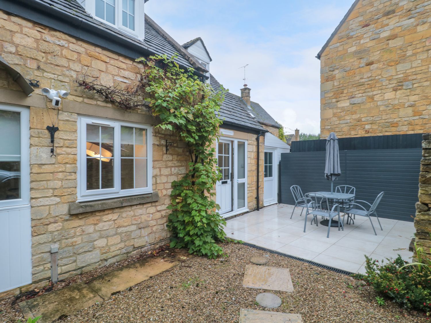 An outdoor patio with stone walls a gravel path a metal table with chairs and an umbrella at 4 Noel Court in Chipping Campden