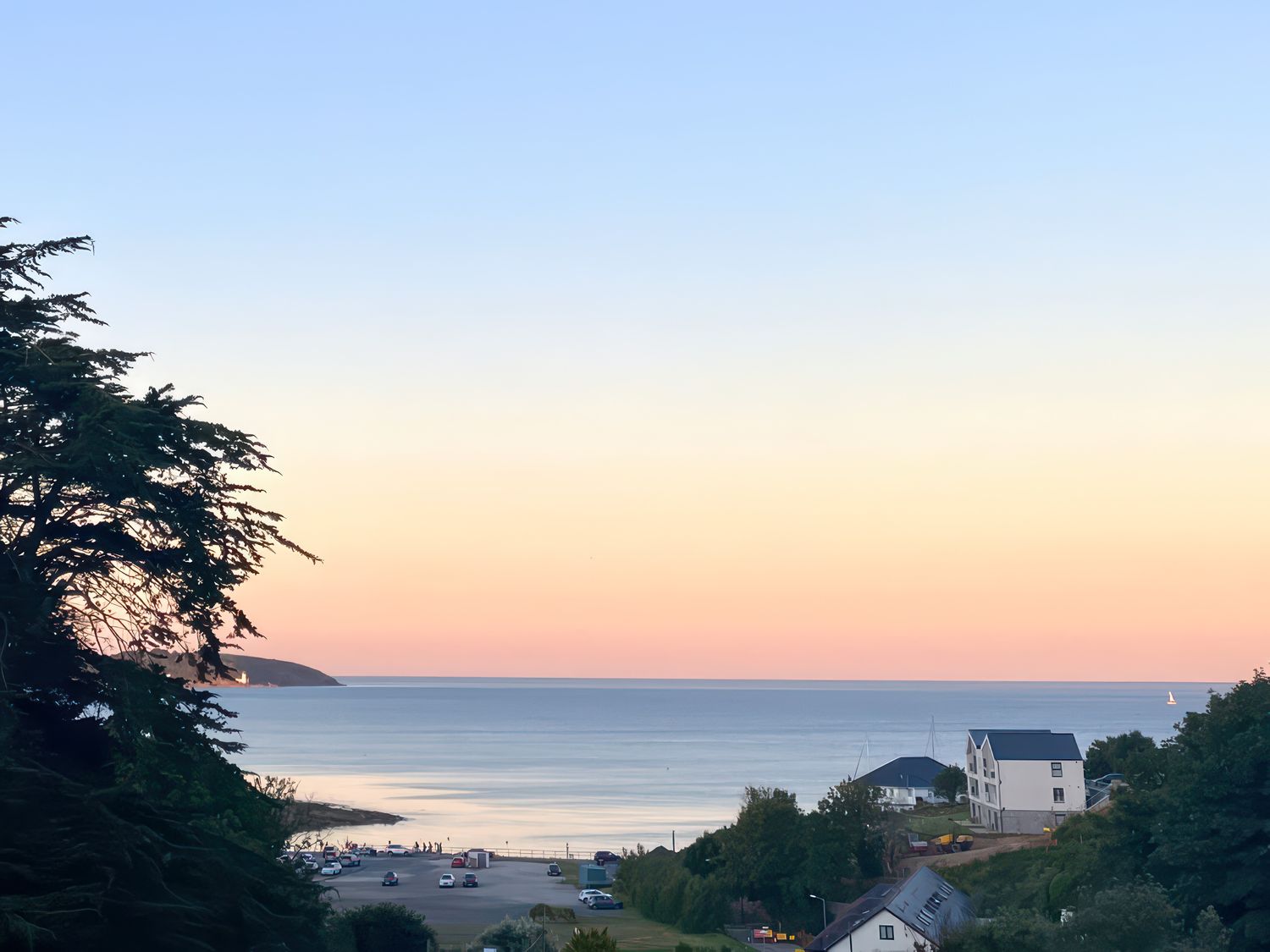 A coastal view with buildings, parked cars, trees, and a calm sea at sunset at Sea Dream in Falmouth