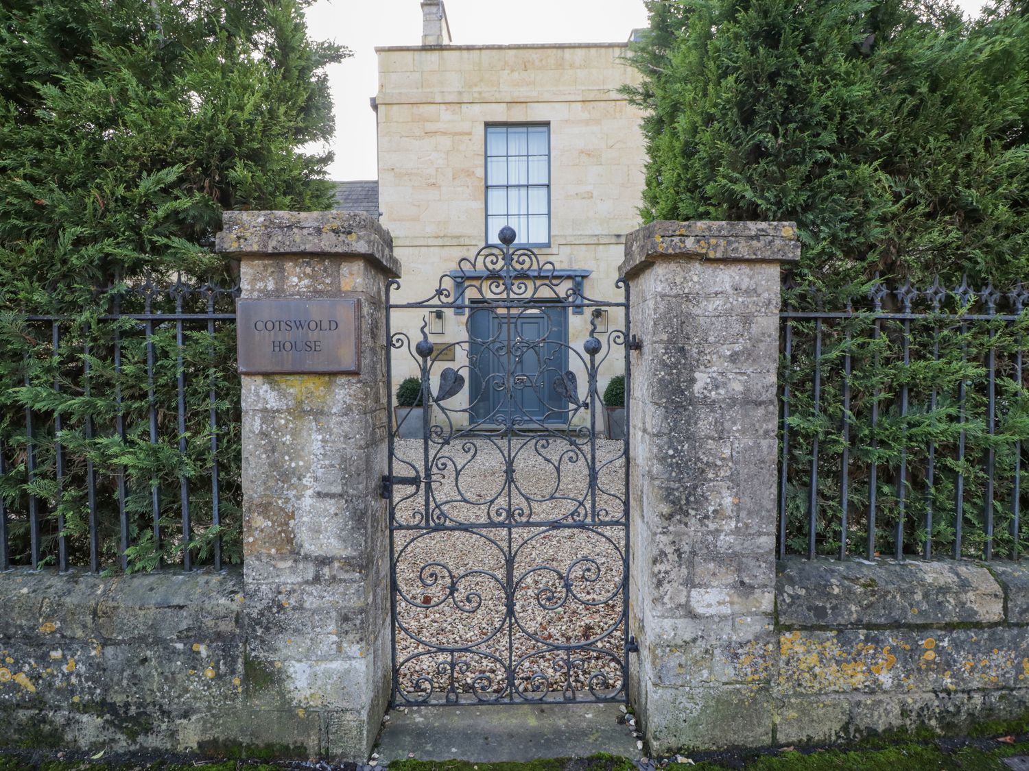 An iron gate between stone pillars with a sign reading Cotswold House and a stone building behind in Moreton-In-Marsh