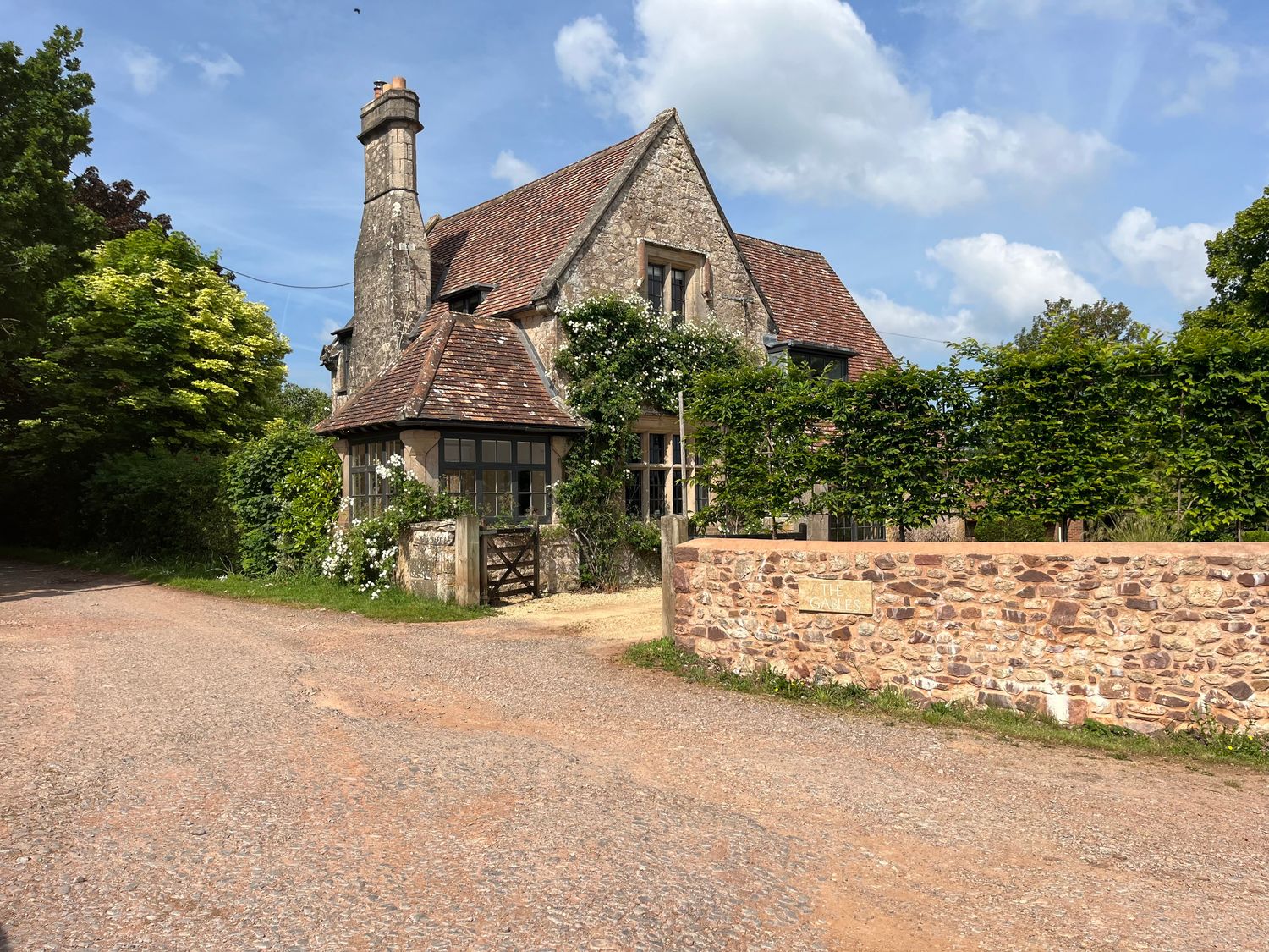 A stone house with a tiled roof surrounded by greenery and a stone wall with a sign that reads The Gables in Spaxton near Nether Stowey
