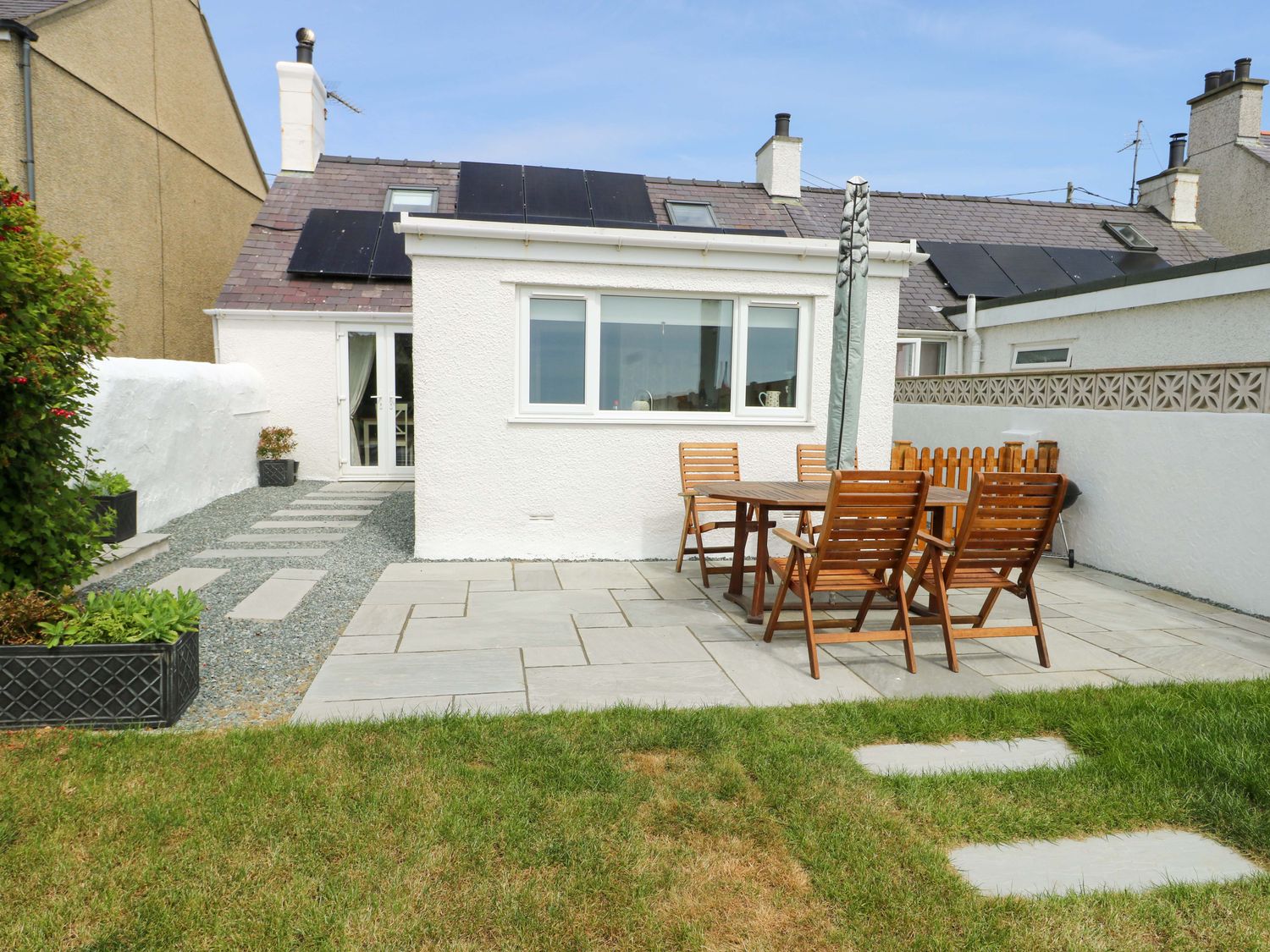 A backyard patio with wooden table and chairs on stone tiles beside a white house at Ty Bach Llanfaelog near Rhosneigr