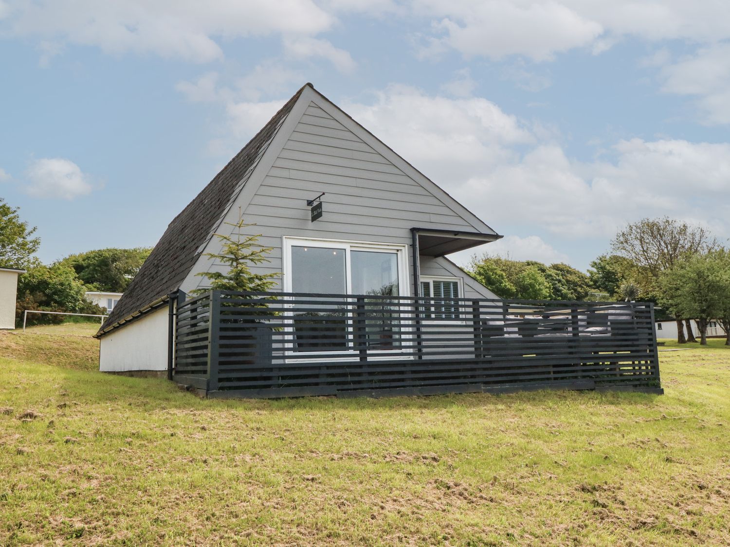 An exterior view of an a-frame house with a black fenced porch on a grassy area at La Bella Vista in Kilkhampton