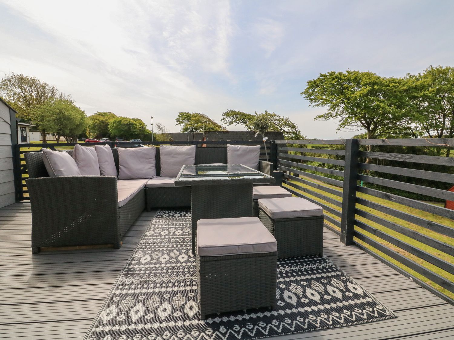 An outdoor deck with a sectional sofa, cushioned stools, a glass table, and a patterned rug at La Bella Vista in Kilkhampton