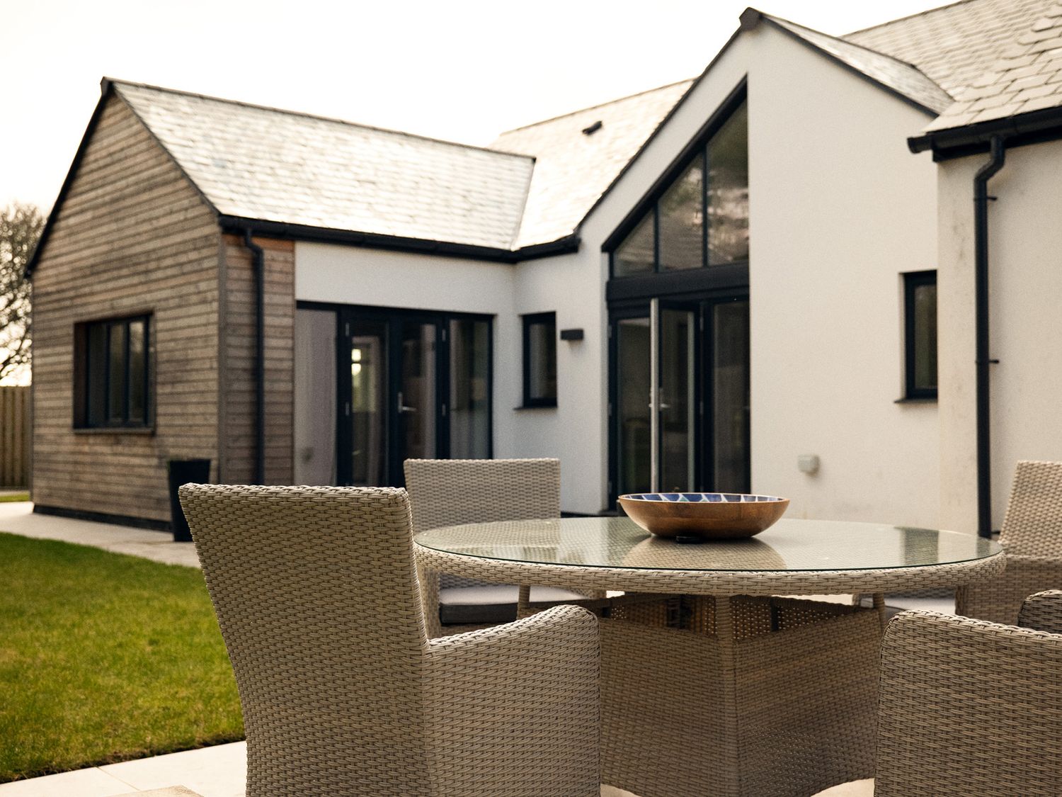 An outdoor patio with a round glass table and wicker chairs in front of a modern house at Kerrek Lodge in Mylor Bridge