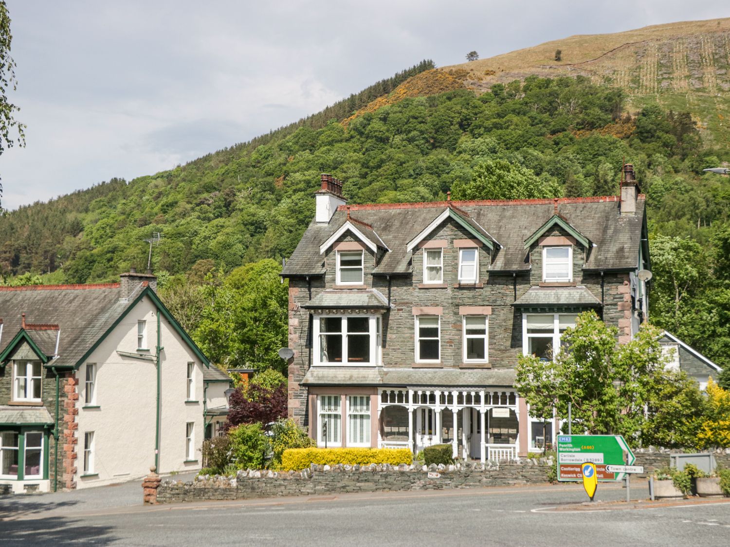 A large stone house with multiple windows and chimneys next to a road with directional signs in front of a forested hill